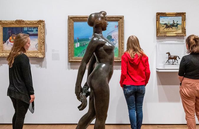 A bronze statue of a nude female form with visitors looking at paintings in the background.