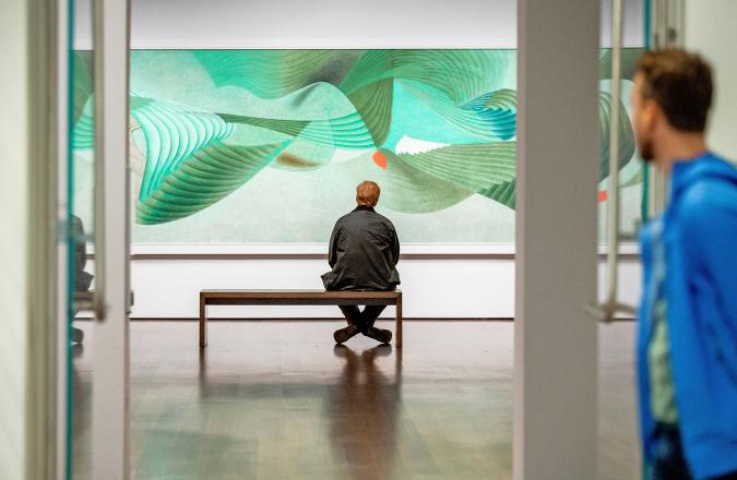 A visitor sits on a bench in front of a large painting with swooping abstract lines in gray, green, and blue. Another visitor looks on in the blurred foreground.