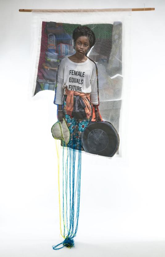 A print on fabric features a Black woman in a white T-shirt with the words “Female equals future.” Blue, yellow, and green threads hang from the bottom of the work. Une impression sur tissu représente une femme noire portant un T-shirt blanc avec les mots « Female equals future ». Des fils bleus, jaunes et verts pendent du bas de l’œuvre.