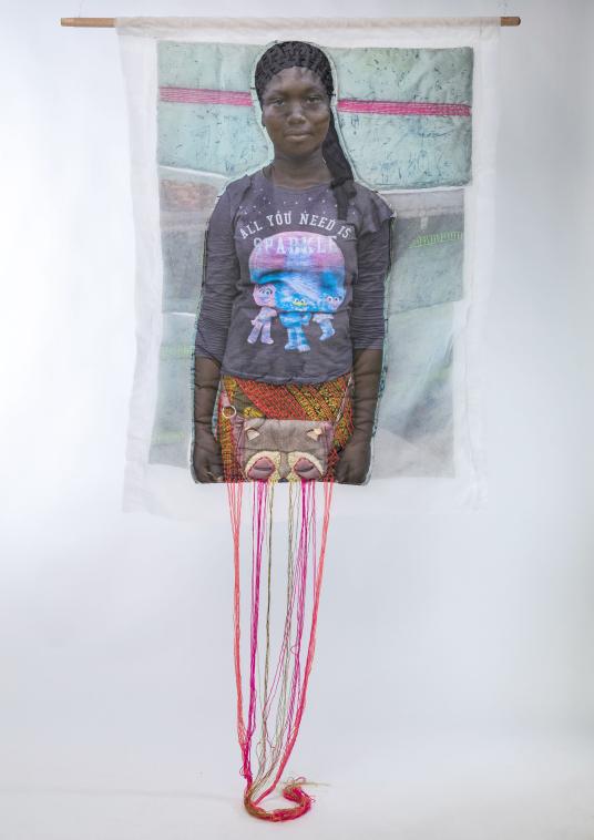 A print on fabric features the image of a Black woman wearing a gray shirt that reads “All you need is sparkle.” Multicolored threads hang from the bottom of the work. Un imprimé sur tissu représente l'image d'une femme noire portant une chemise grise sur laquelle est inscrit « All you need is sparkle ». Des fils multicolores pendent du bas de l'œuvre.