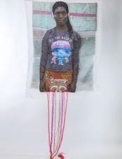 A print on fabric features the image of a Black woman wearing a gray shirt that reads “All you need is sparkle.” Multicolored threads hang from the bottom of the work. Un imprimé sur tissu représente l'image d'une femme noire portant une chemise grise sur laquelle est inscrit « All you need is sparkle ». Des fils multicolores pendent du bas de l'œuvre.