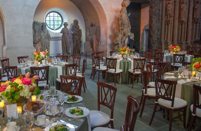 A hall with a rustic tile floor is setup for dinner service and surrounded by statues.