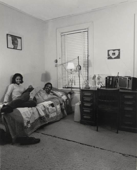Portrait of a young couple smiling casually on a bed in a carpeted room beside a desk and chair.