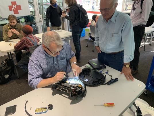 A man holds a tool over an opened-up device while another man looks on.