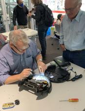 A man holds a tool over an opened-up device while another man looks on.