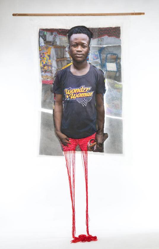 A print on fabric features a Black man in a black T-shirt with the words “Wonder Woman.” Red threads hang from the bottom of the work. Une impression sur tissu représente un homme noir portant un T-shirt noir avec l’inscription « Wonder Woman ». Des fils rouges pendent du bas de l'œuvre.
