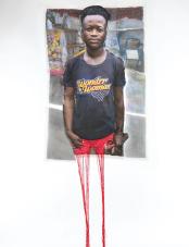 A print on fabric features a Black man in a black T-shirt with the words “Wonder Woman.” Red threads hang from the bottom of the work. Une impression sur tissu représente un homme noir portant un T-shirt noir avec l’inscription « Wonder Woman ». Des fils rouges pendent du bas de l'œuvre.