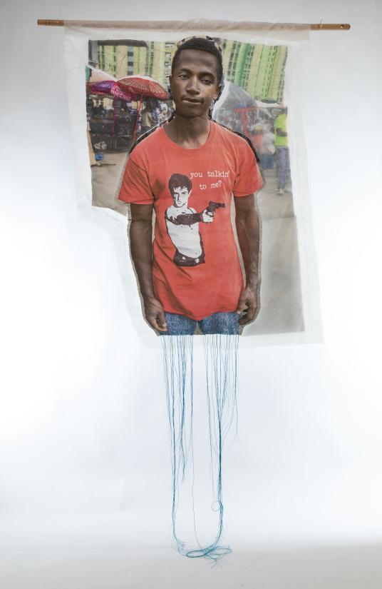 A photographic print on fabric features a Black man wearing a red T-shirt with text that reads “You talkin’ to me?.” Blue threads hang from the bottom of the work. Une impression photographique sur tissu représente un homme noir portant un T-shirt rouge avec le texte « You talkin’ to me? ». Des fils bleus pendent du bas de l’œuvre.
