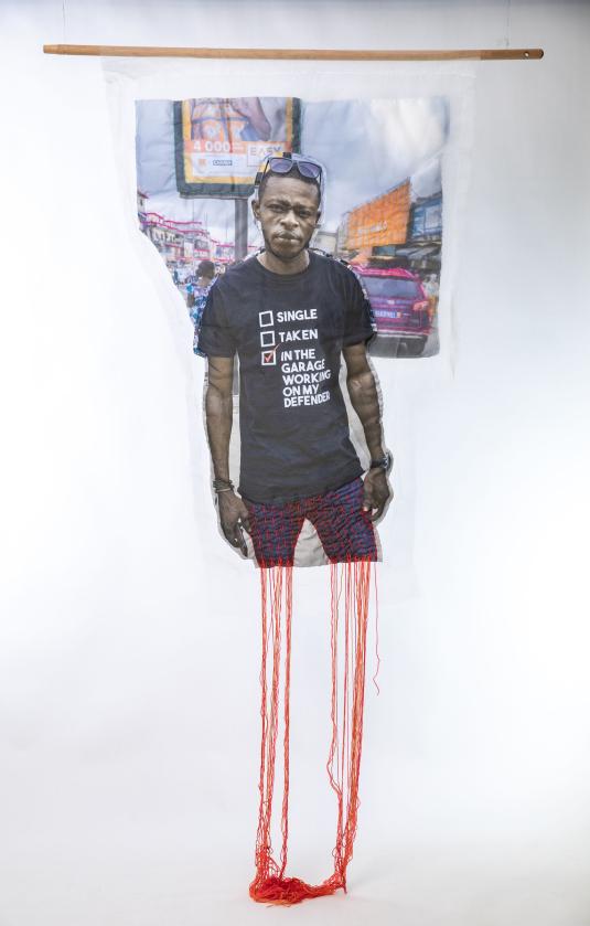 A print on white fabric features a Black man in a black shirt with white text. Red and orange threads hang from the bottom of the work. Un imprimé sur tissu blanc représente un homme noir portant une chemise noire avec du texte blanc. Des fils rouges et oranges pendent du bas de l'œuvre.