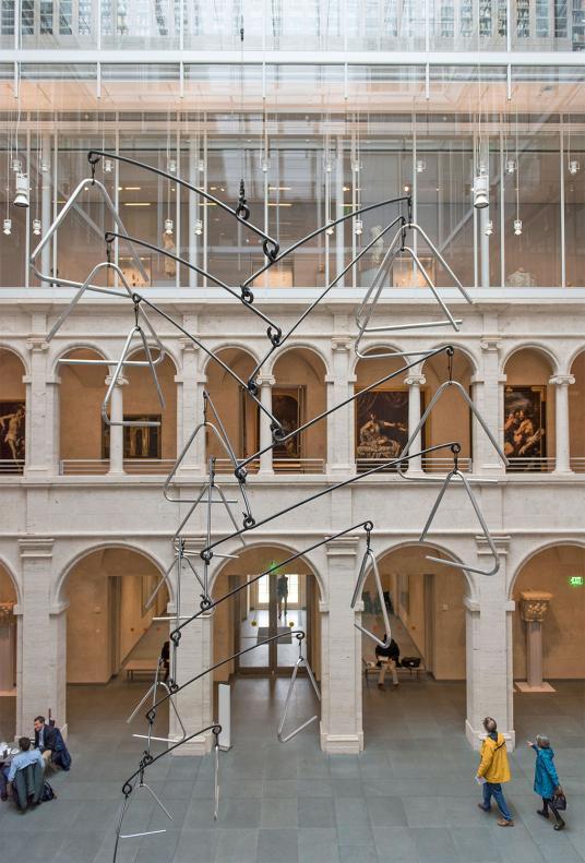 A view from above of one side of a courtyard with three levels. The bottom two levels have arches, and a few people can be seen in the main floor of the courtyard. A large metal sculpture consisting of several interlocking triangles hangs from the ceiling.