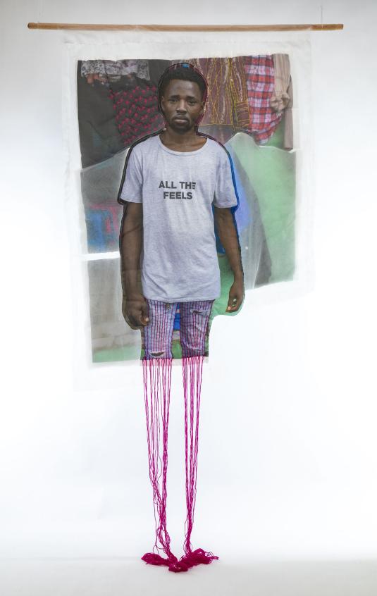 A print on white fabric features a Black man in a white T-shirt that reads “All the feels.” Magenta embroidery hangs at the bottom of the work. Une impression sur tissu blanc représente un homme noir portant un t-shirt blanc avec l'inscription « All the feels ». Des broderies magenta pendent au bas de l'œuvre.