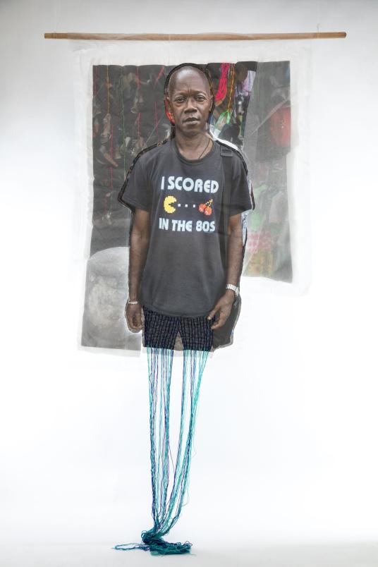 A print on fabric features the image of a Black man wearing a black shirt that reads “I scored in the 80s.” Teal and purple threads hang from the bottom of the work. Une impression sur tissu représente un homme noir portant un T-shirt noir avec l'inscription « I scored in the 80s ». Des fils turquoise et violets pendent au bas de l’œuvre.