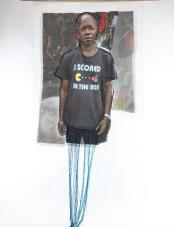 A print on fabric features the image of a Black man wearing a black shirt that reads “I scored in the 80s.” Teal and purple threads hang from the bottom of the work.  Une impression sur tissu représente un homme noir portant un T-shirt noir avec l'inscription « I scored in the 80s ». Des fils turquoise et violets pendent au bas de l’œuvre.
