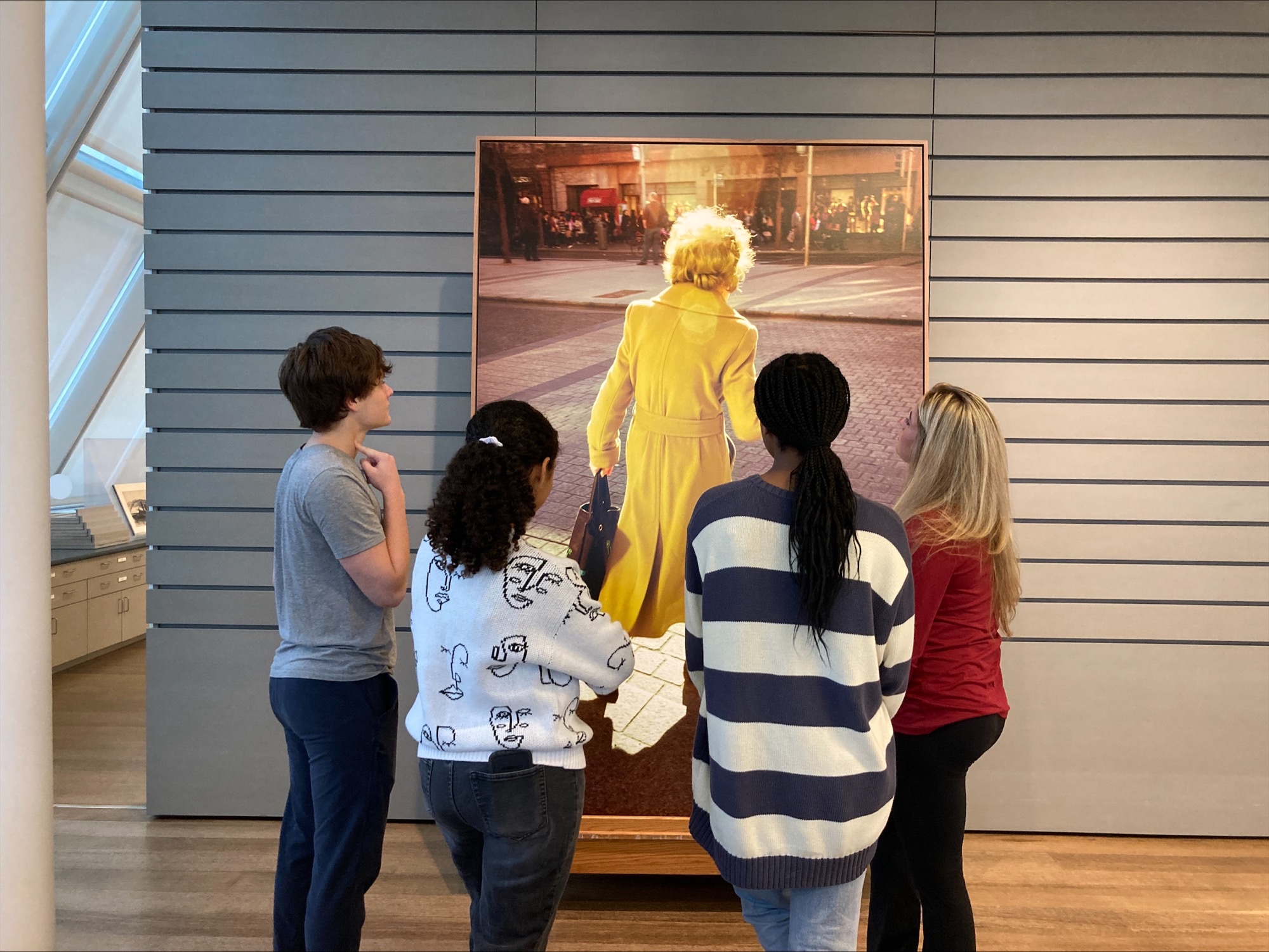 Four students, with their backs to the camera, look at a large rectangular photograph of an older woman in a yellow coat, walking across a city street.
