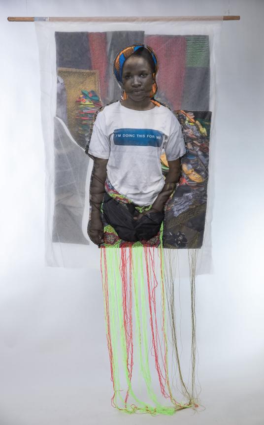 A photographic print on fabric features a Black woman wearing a colorful head covering, a white T-shirt that reads “I’m doinc this for me,” and a red and green embroidered skirt. Un tirage photographique sur tissu représente une femme noire portant un couvre-chef coloré, un T-shirt blanc avec l'inscription « I’m doinc this for me » et une jupe brodée rouge et verte.