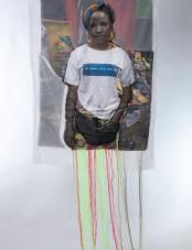 A photographic print on fabric features a Black woman wearing a colorful head covering, a white T-shirt that reads “I’m doinc this for me,” and a red and green embroidered skirt. Un tirage photographique sur tissu représente une femme noire portant un couvre-chef coloré, un T-shirt blanc avec l'inscription « I’m doinc this for me » et une jupe brodée rouge et verte.