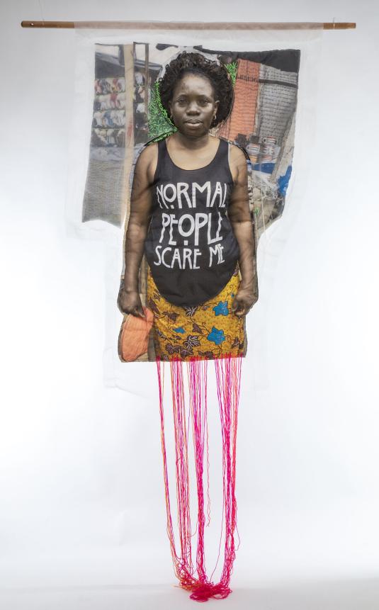 A print on white fabric features a Black woman in a black tank top that reads “Normal people scare me.” Pink, red, and orange threads hang from the bottom of the work.  Une impression sur tissu blanc représente une femme noire portant un débardeur noir avec l'inscription « Normal people scare me ». Des fils roses, rouges et orange pendent au bas de l'œuvre.