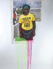 A print on fabric features a Black woman in a yellow T-shirt with the words “Touch me and your first boxing lesson is free.” Green, yellow, and pink threads hang from the bottom of the work. Une impression sur tissu représente une femme noire portant un T-shirt jaune avec l’inscription « Touch me and your first boxing lesson is free ». Des fils verts, jaunes et roses pendent au bas de l’œuvre.