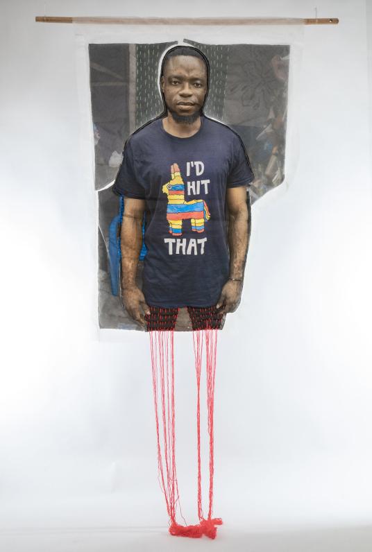 A print on fabric features the image of a Black man wearing a black shirt that reads “I’d hit that.” Red threads hang from the bottom of the work. Un motif sur tissu représente l’image d’un homme noir portant un t-shirt noir avec l’inscription « I’d hit that ». Des fils rouges pendent du bas de l’œuvre.