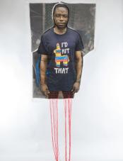 A print on fabric features the image of a Black man wearing a black shirt that reads “I’d hit that.” Red threads hang from the bottom of the work. Un motif sur tissu représente l’image d’un homme noir portant un t-shirt noir avec l’inscription « I’d hit that ». Des fils rouges pendent du bas de l’œuvre.