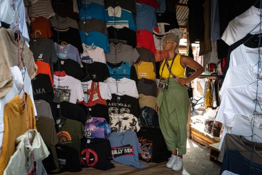 Photograph of a Black woman standing in front of T-shirts hanging on a wall. Photographie d’une femme noire debout devant des T-shirts accrochés à un mur.