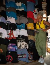 Photograph of a Black woman standing in front of T-shirts hanging on a wall. Photographie d’une femme noire debout devant des T-shirts accrochés à un mur.