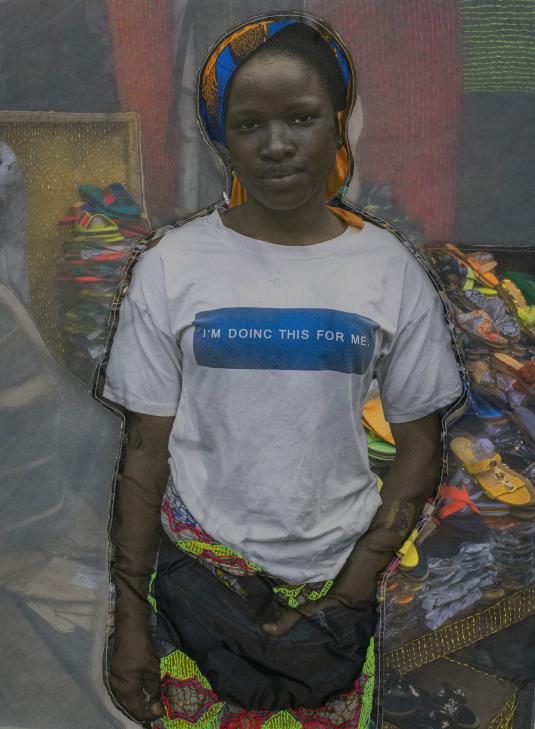 A photographic print on fabric features a Black woman wearing a colorful head covering, a white T-shirt that reads “I’m doinc this for me,” and a red and green embroidered skirt. Une impression photographique sur du tissu blanc montre une femme noire pourtant un couvre-chef bleu et orange, un T-shirt blanc sur lequel est inscrit « I’m doinc this for me » et une jupe brodée de fils rouges et vert fluo.