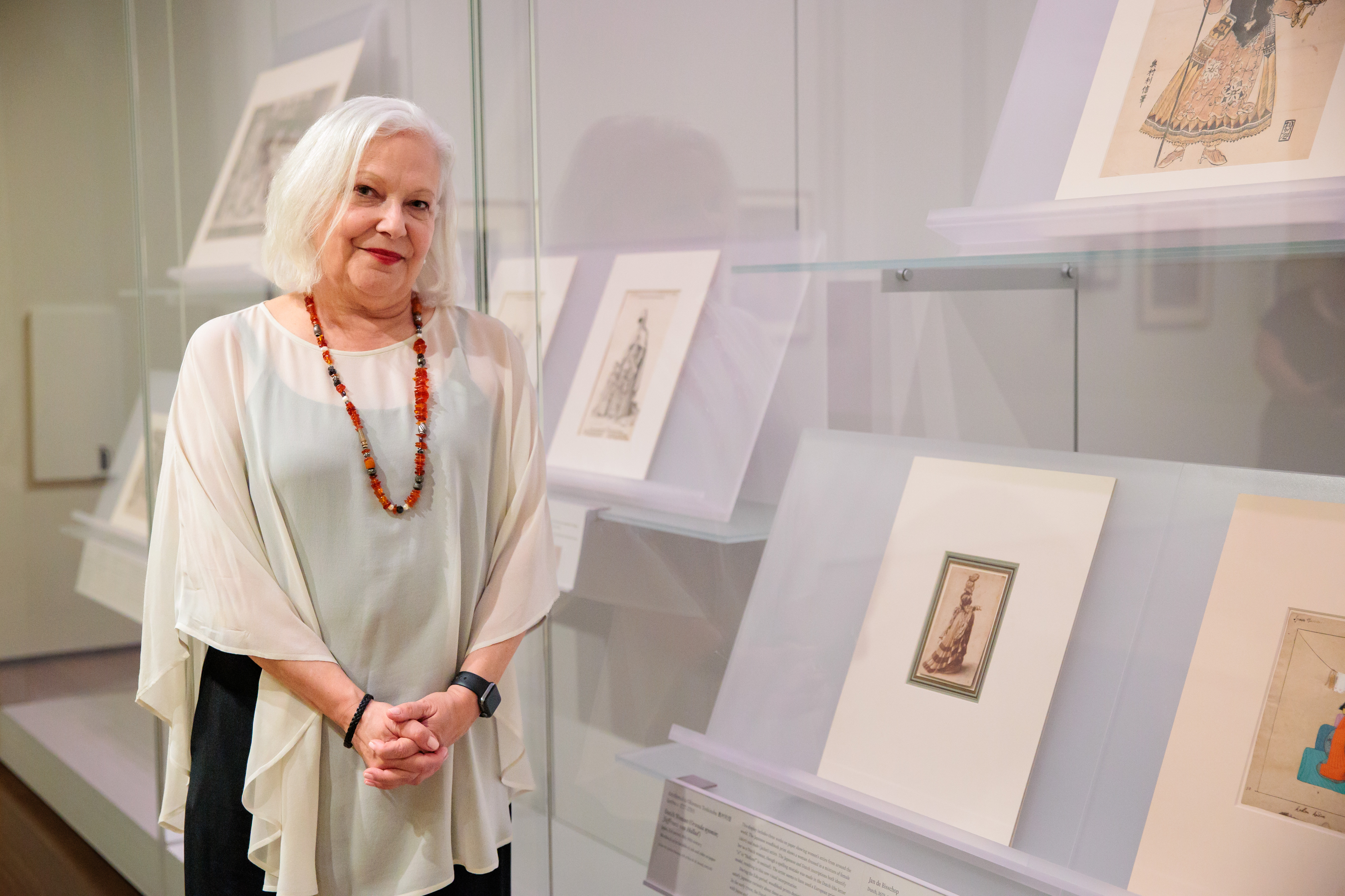 A woman in a white shawl standing next to a drawing displayed within a glass case.